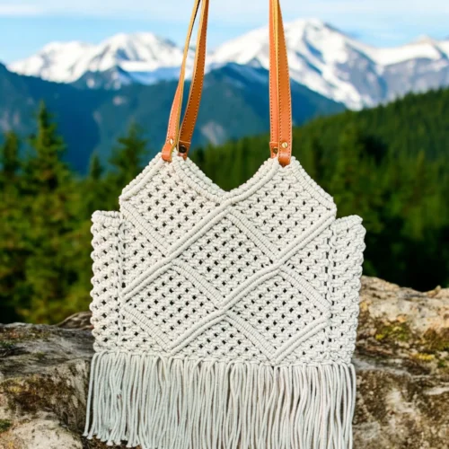 Handwoven white macramé bag with tan leather straps and fringe detailing, displayed against a scenic mountain backdrop.