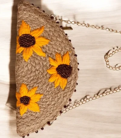 Make a sunny statement with the ATICUE half moon jute clutch. Features hand-embroidered sunflowers, zipper closure, and gold shoulder chain for effortless charm.